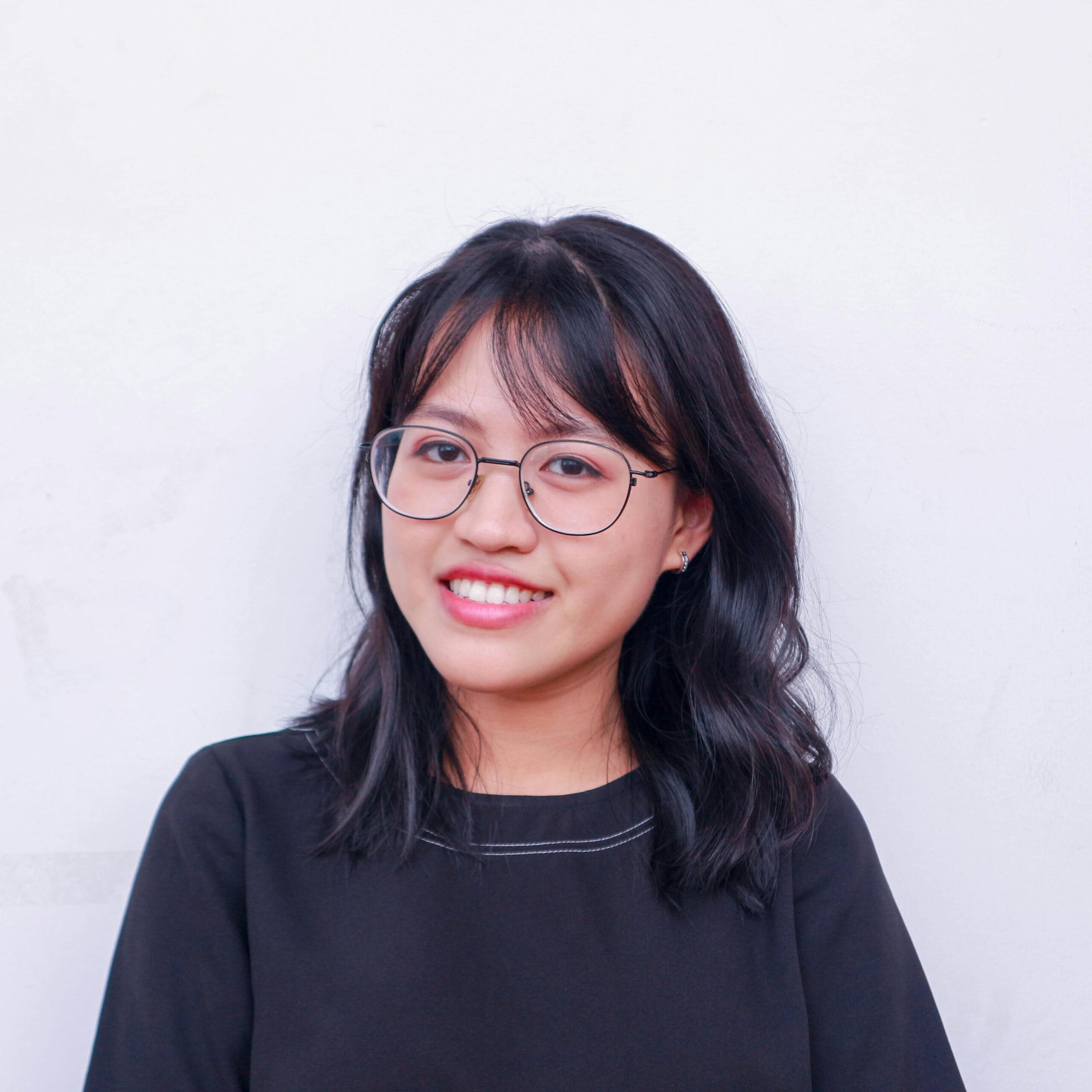 Home 13 Portrait of a young woman with glasses and black hair smiling against a plain background.